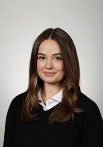 Headshot portrait of young brunette woman in white-collared shirt and black sweater, gray background
