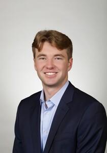 Headshot photograph of smiling young man with light brown hair wearing navy blazer